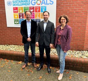 Drei Personen stehen vor einer Tafel mit den Nachhaltigkeitszielen der Agenda 2030, die an einer Außenwand eines Gebäudes hängt.
