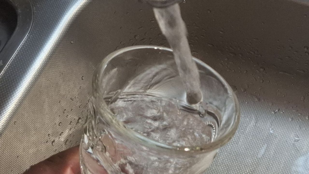 Das Bild zeigt eine Hand mit einem Wasserglas, das in einer Spüle mit Trinkwasser gefüllt wird.