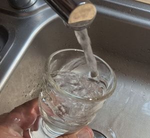 Das Bild zeigt eine Hand mit einem Wasserglas, das in einer Spüle mit Trinkwasser gefüllt wird.
