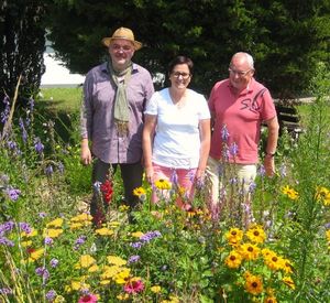 Auf dem Bild sind drei Personen hinter einer bunten Blühwiese und vor zwei grünen Bäumen zu erkennen.
