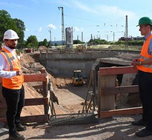 Auf dem Bild sind zwei Männer zu sehen, welche mit einer Wahnweste und einem Schutzhelm ausgestattet sind. Sie stehen vor einer Treppe, die zu einer Baustelle führt.