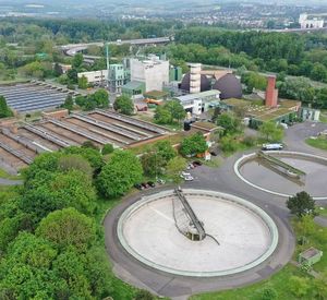 Auf dem Bild sind zwei große runde Becken und fabrikartige Gebäude der Kläranlage zu sehen. Die Anlage wird von Bäumen umrandet.