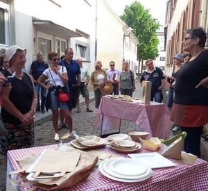 Das Bild zeigt eine Gruppe von Menschen, die auf einer kleinen Straße um zwei Tische herum stehen und diese betrachten. Auf dem einen Tisch befinden sich Wegwerfgeschirr, Papiertaschen und weitere Verpackungen.