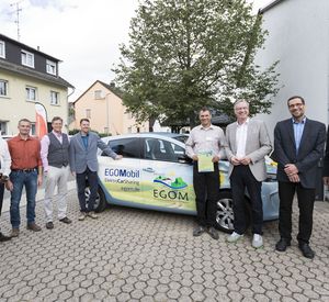 Das Bild zeigt neun Personen, die vor einem Carsharingauto stehen.