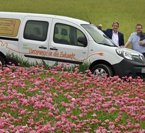 Das Foto zeigt eine Blumenwiese und ein Elektroauto, hinter dem zwei Männer stehen.