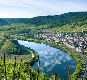 Das Foto zeigt den Blick von einem Berg auf einen Fluss mit einer naheligenden Ortschaft. Zudem sind Weinberge und bewaldete Hügel zu erkennen. Das Wetter ist sehr sonnig und die wenigen Wolken spiegeln sich im Fluss. 